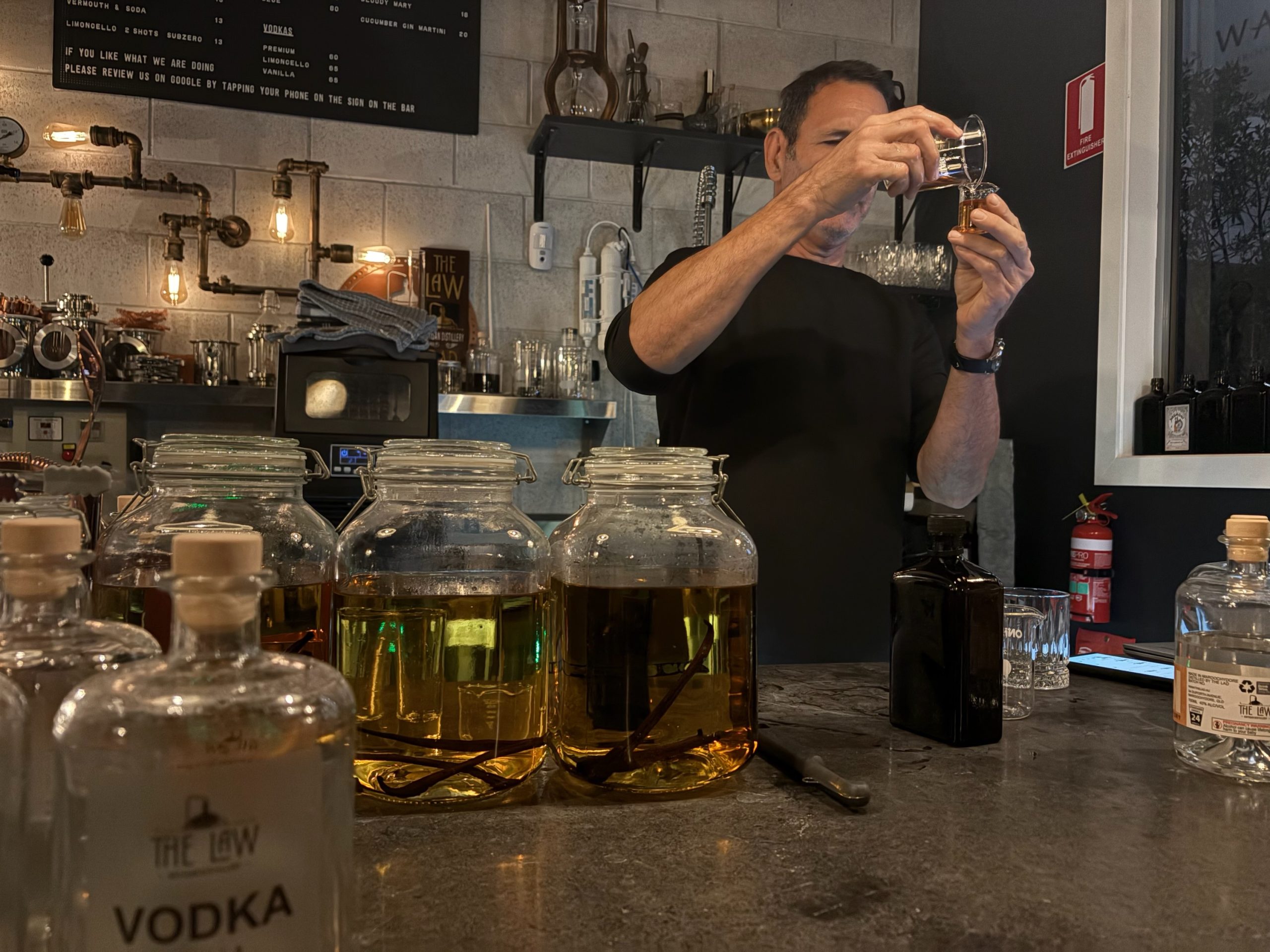 The distiller pouring a measure at the bar of The Law Artisan Distillery, with jars of vanilla infusing in the foreground.